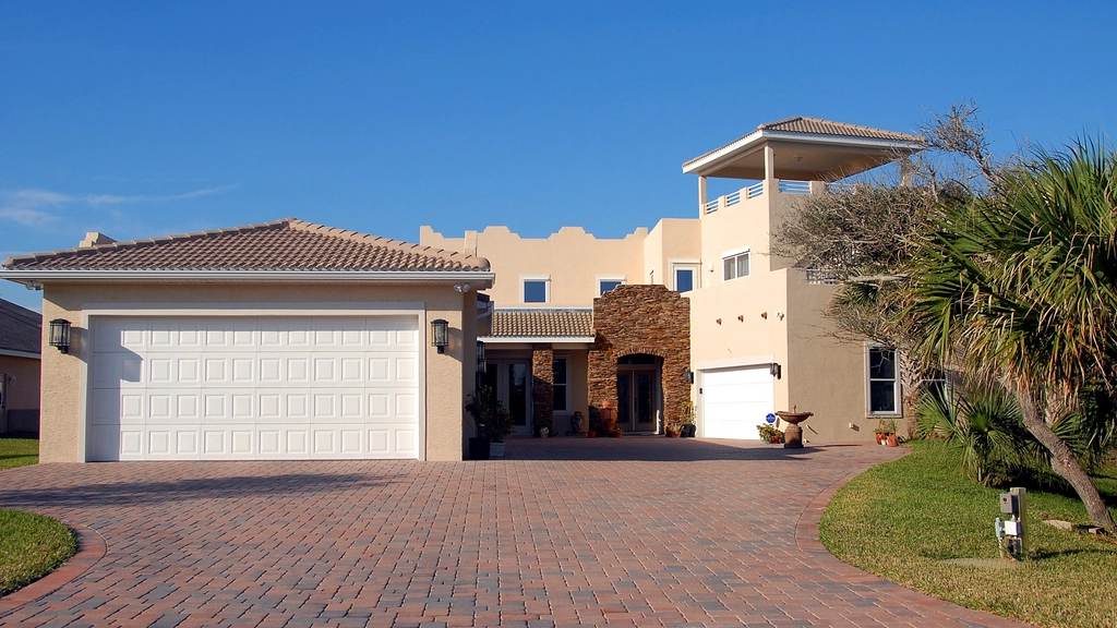Red brick paver driveway leading to a tan stucco home with palm trees